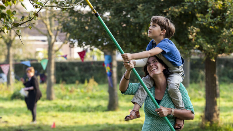 Een vrouw draagt een kind op haar schouders; samen plukken ze fruit met een stok in een grasveld.