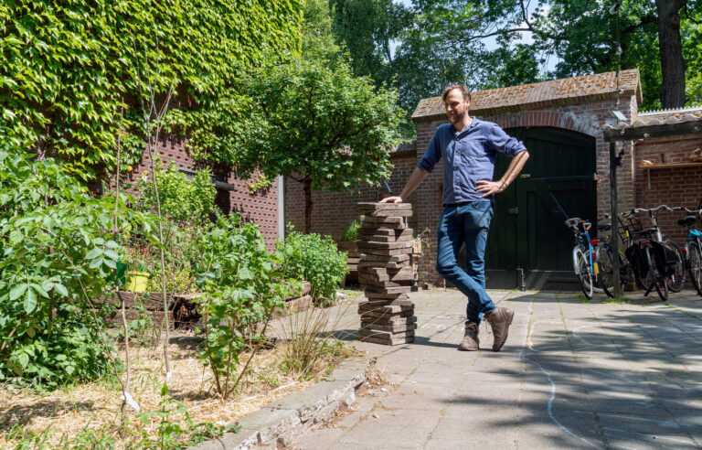 Persoon leunt op stapel stenen in zonnige tuin met begroeiing en fietsen tegen oude muur.