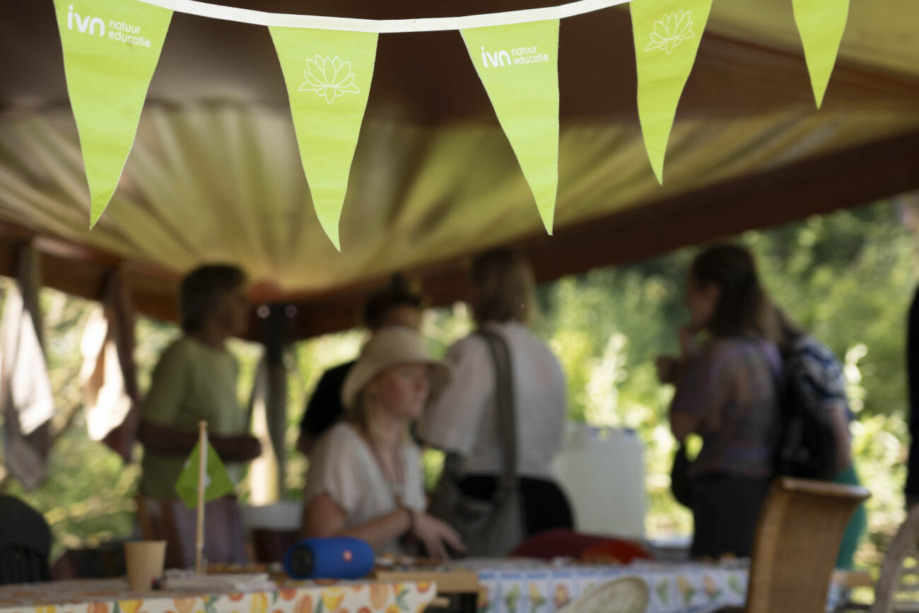 Groene vlaggetjes met IVN-logo boven tafels in een tent met mensen in gesprek.
