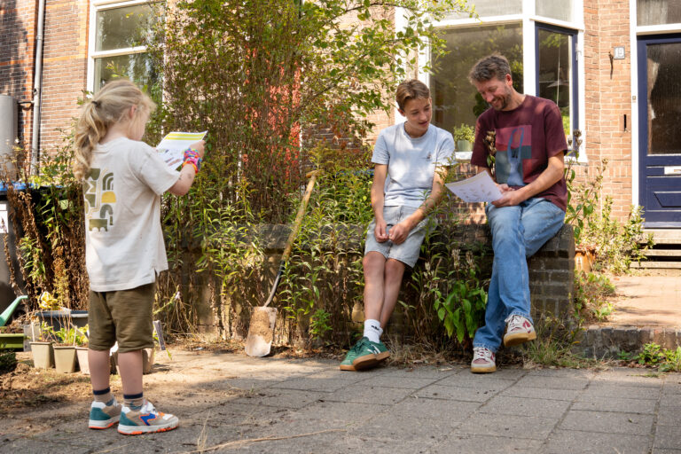 Kind leest boek voor aan twee zittende mensen in een tuin met planten en schoppen.