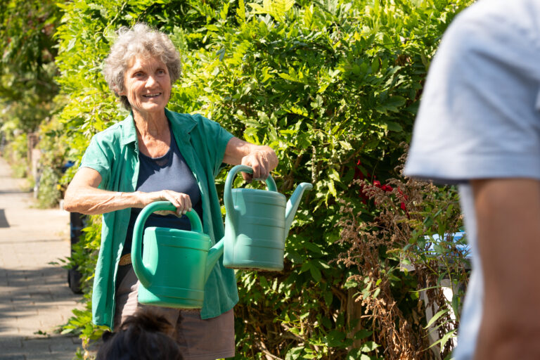 Een vrouw met grijs haar draagt twee groene gieters in een groene tuin.