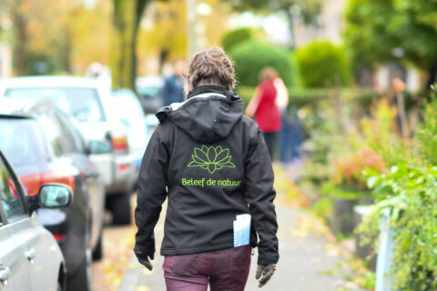 Persoon loopt buiten op stoep met zwarte jas met groene lotusbloem en tekst "Beleef de natuur".