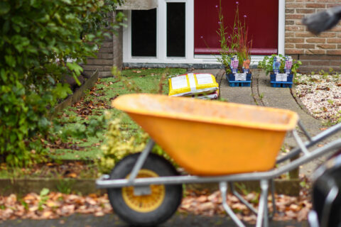 Een gele kruiwagen voor een huis met plantenbakken en een zak aarde bij de voordeur.