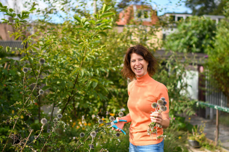 Vrouw in oranje trui glimlacht terwijl ze bloemen knipt in een groene tuin.