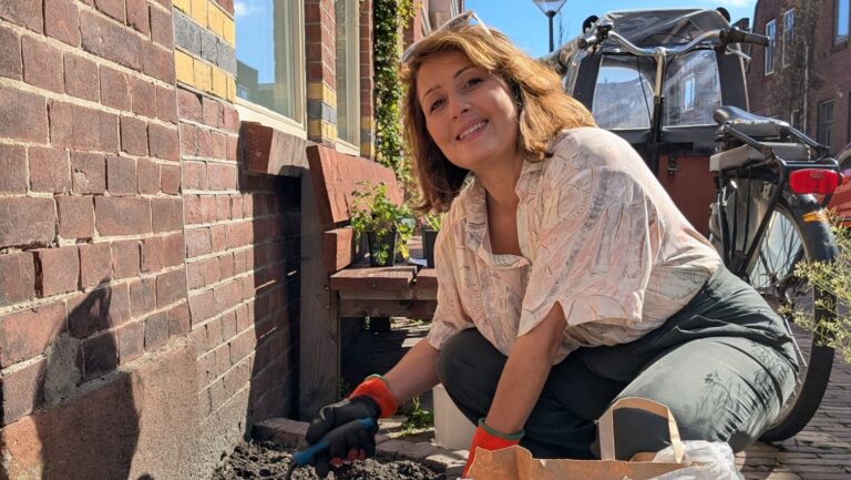 Een vrouw knielt buiten bij een baksteenmuur, lachend en tuinierend naast een fiets.