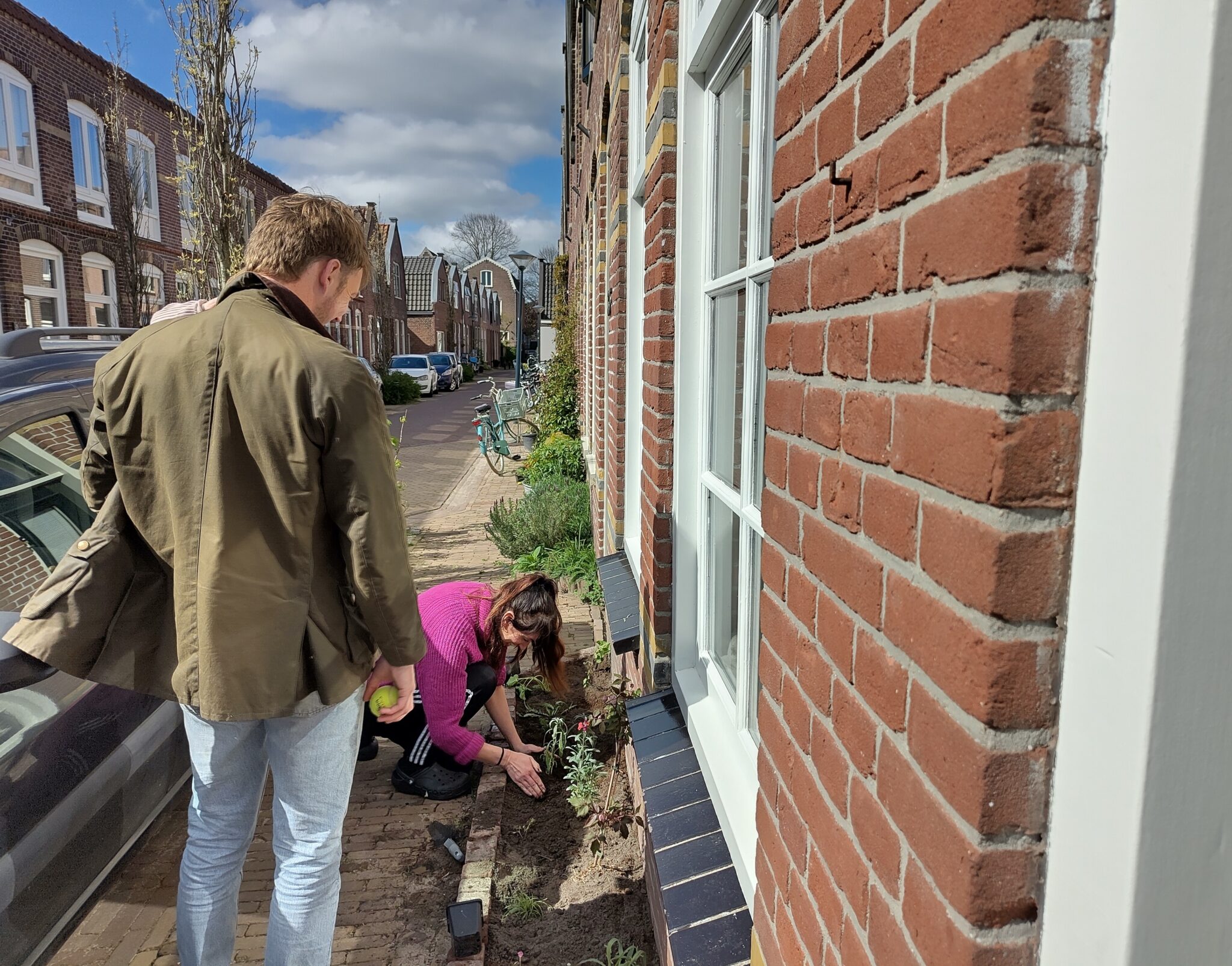 Man en vrouw planten bloemen naast huis in zonnige straat met bakstenen gebouwen.