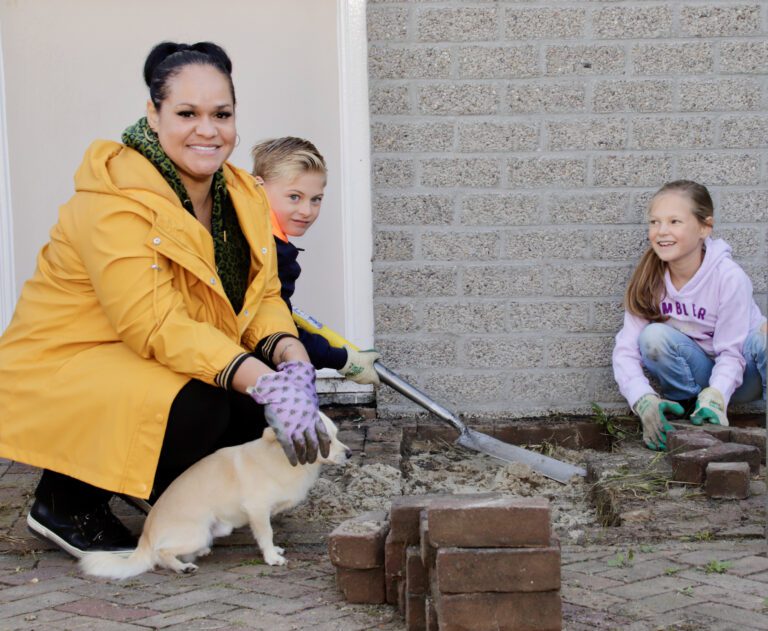 Vrouw en kinderen met hond leggen stenen naast een muur. Ze dragen tuinhandschoenen.