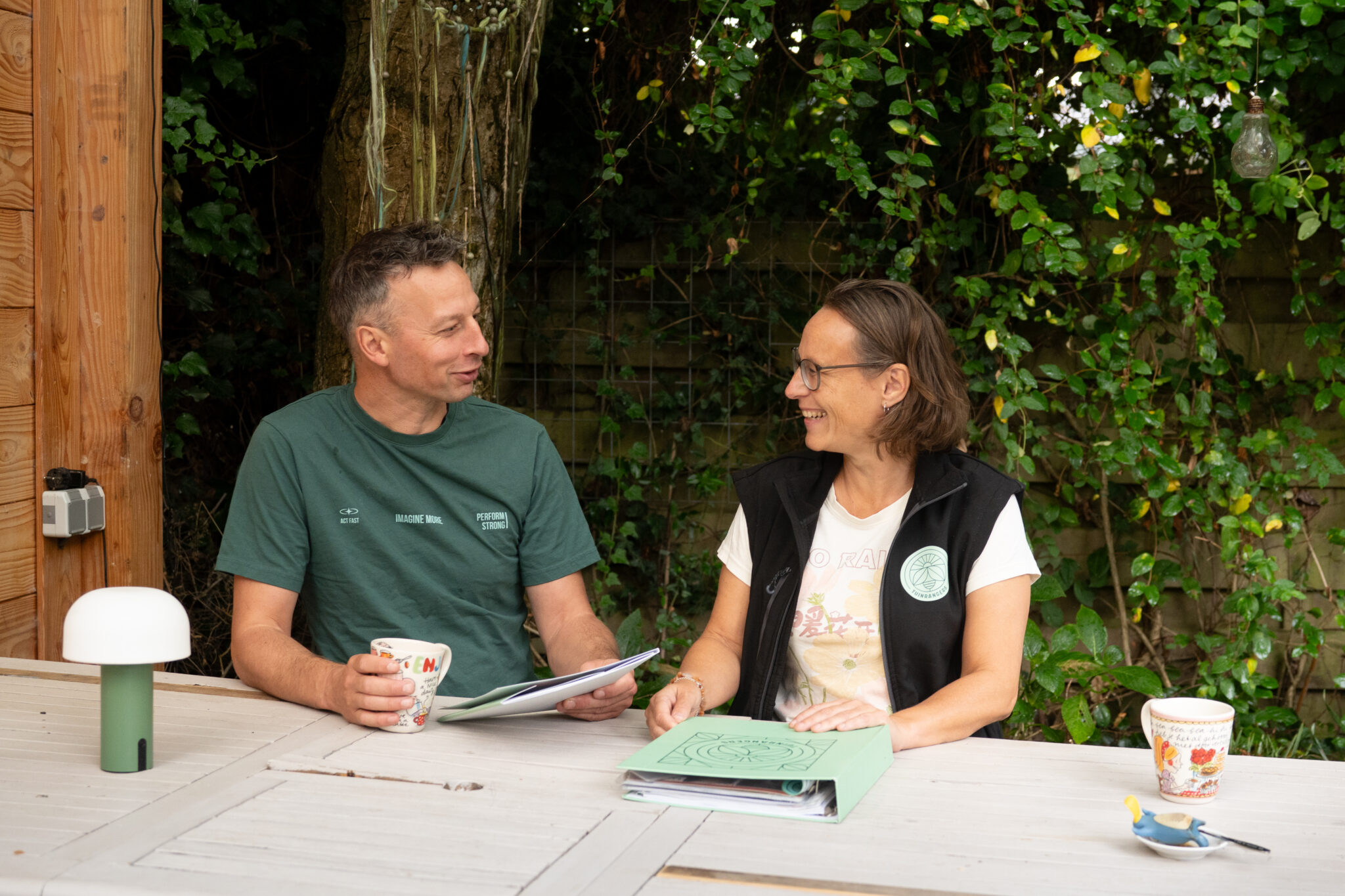 Twee mensen praten en lachen aan een tafel met koffiekopjes en papieren, buiten met een groene achtergrond.