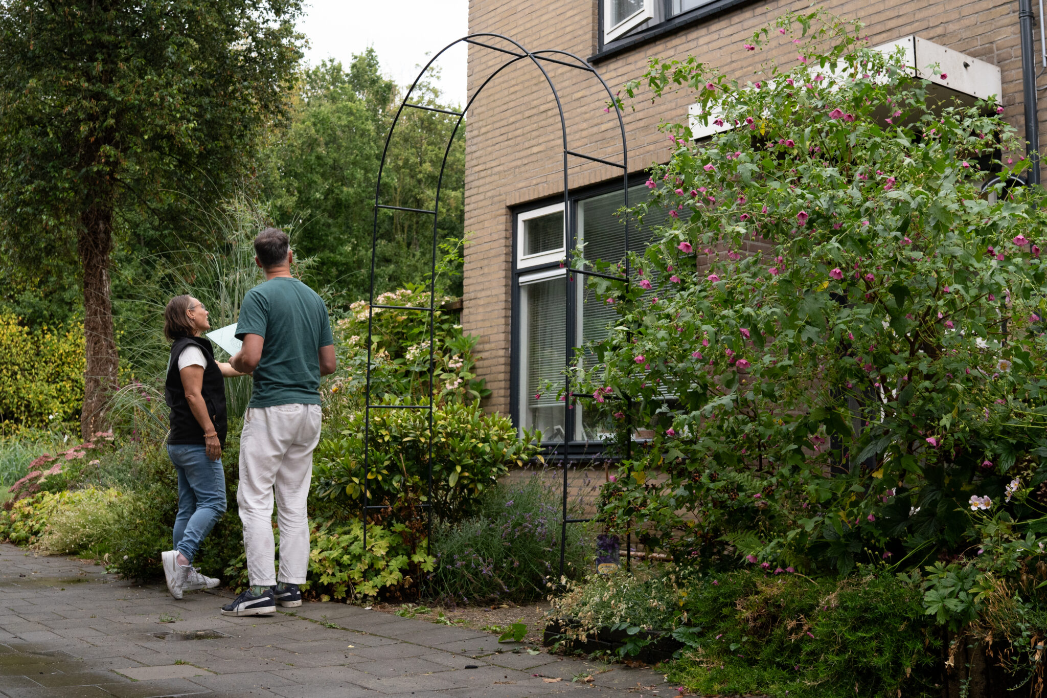Twee mensen staan bij een boog in een groene tuin naast een bakstenen huis.