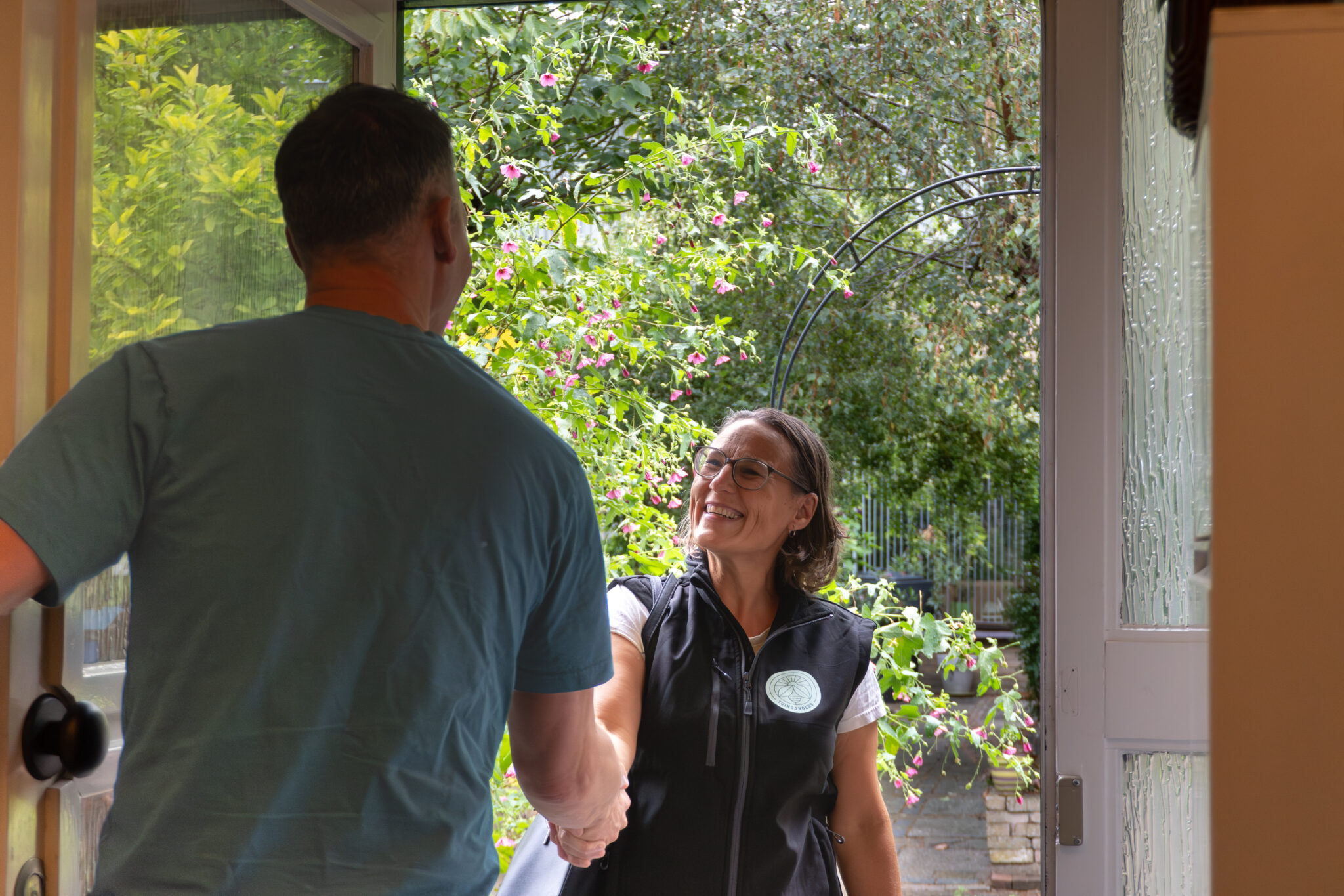 Een vrouw schudt glimlachend de hand van een man bij een deuropening, met groene tuin op achtergrond.