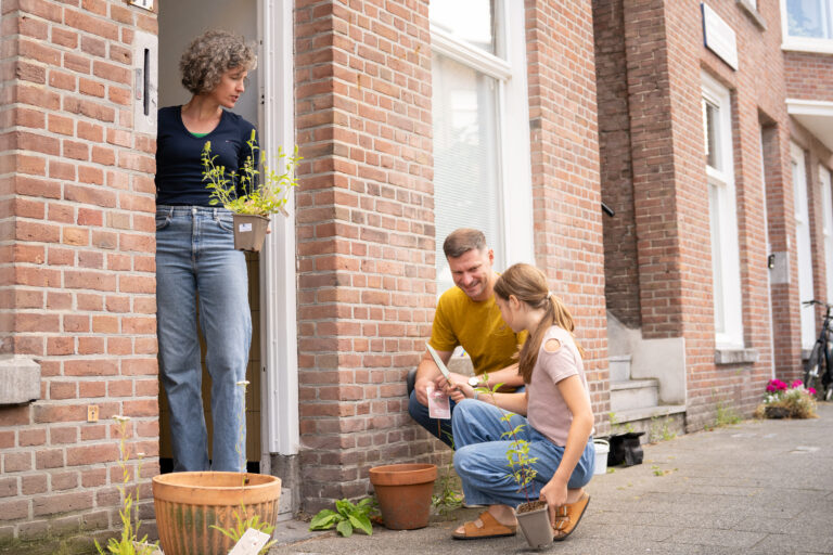 Persoon met plant bij deur kijkt naar man en kind die samen planten in potten plaatsen op stoep.