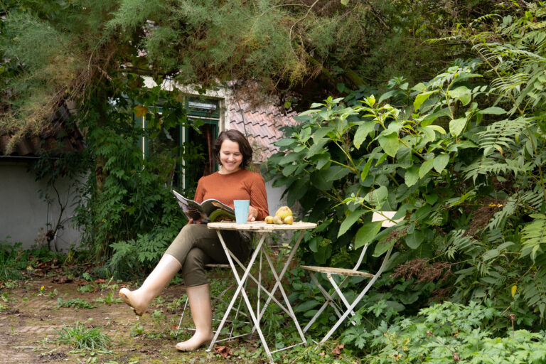 Vrouw leest een tijdschrift aan een tuintafel omringd door groene planten.