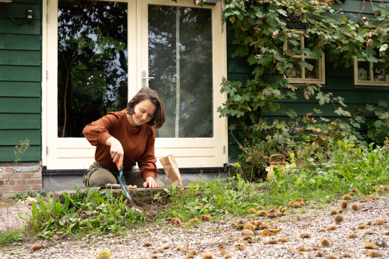 Vrouw tuiniert voor een huis met groene gevel en glazen deuren, omgeven door planten.