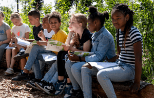 Kinderen lezen en schrijven buiten, zittend op houten blokken, omringd door groen.