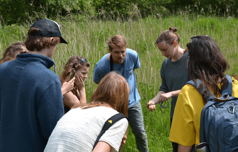 Groep mensen verzameld in een veld, kijken naar iemand die een kleine slang vasthoudt.
