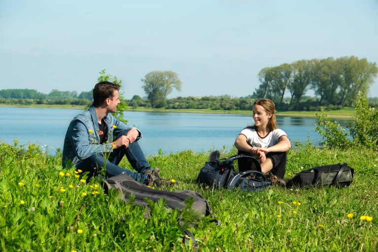 Twee mensen zitten in gras bij een meer, omringd door rugzakken en bloemen, onder een zonnige hemel.