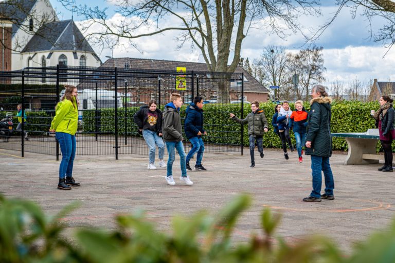 Kinderen spelen op een schoolplein; twee volwassenen kijken toe. In de achtergrond staat een kerk.