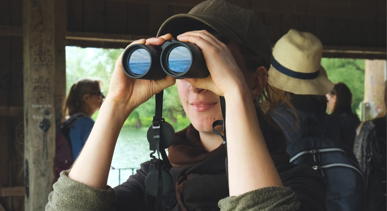Persoon kijkt door een verrekijker, met groen landschap op de achtergrond in een houten schuilplaats.