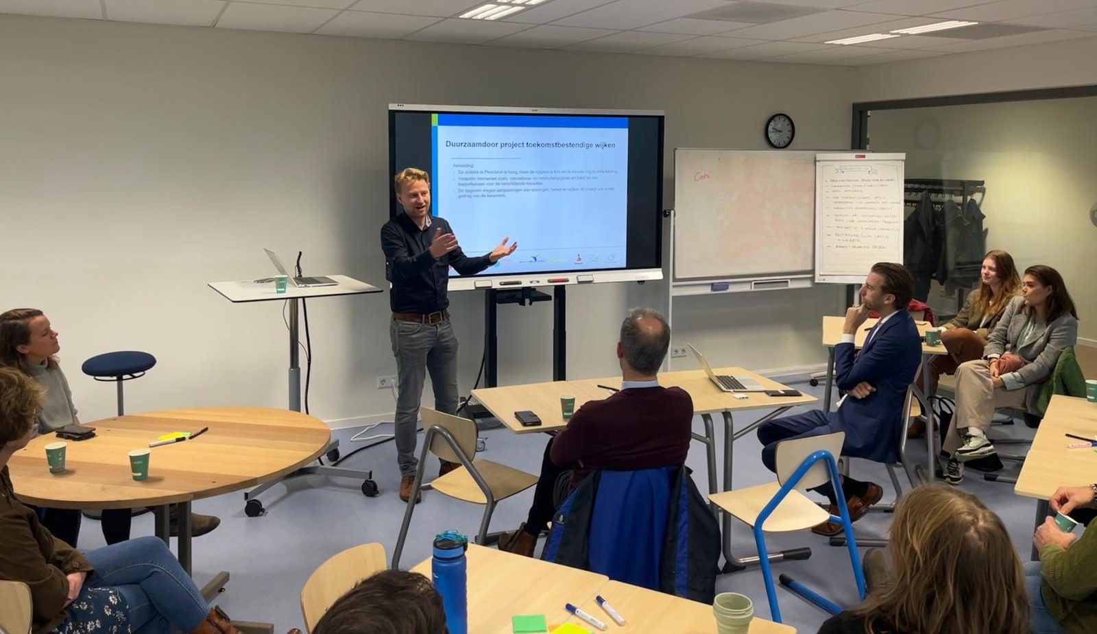 Man geeft presentatie voor een groep in een vergaderruimte met schermpresentatie op achtergrond.