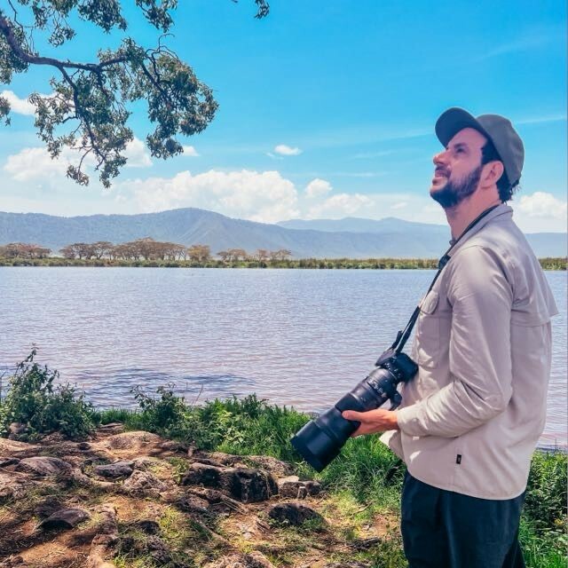 Man met camera kijkt naar boom bij meer met bergen op de achtergrond.