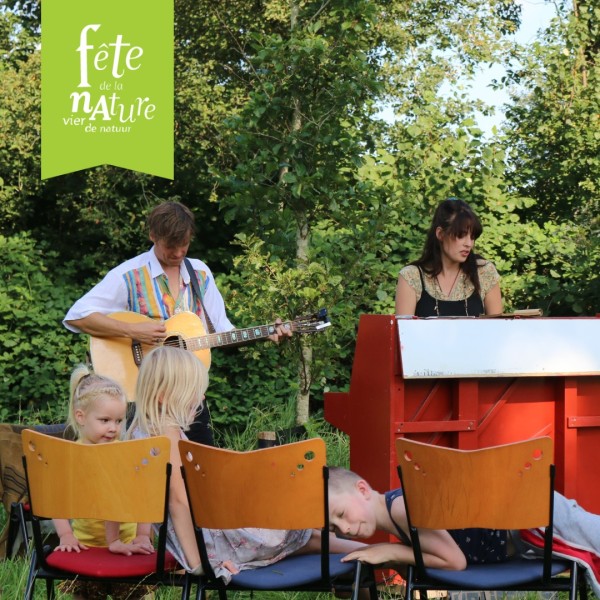 Muzikanten spelen buiten, kinderen zitten op stoelen. Tekst: "Fête de la Nature, vier de natuur".