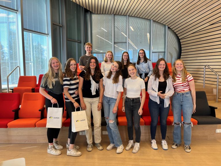 Groep jongeren poseert in een moderne ruimte met rode banken; twee dragen tassen met logo.