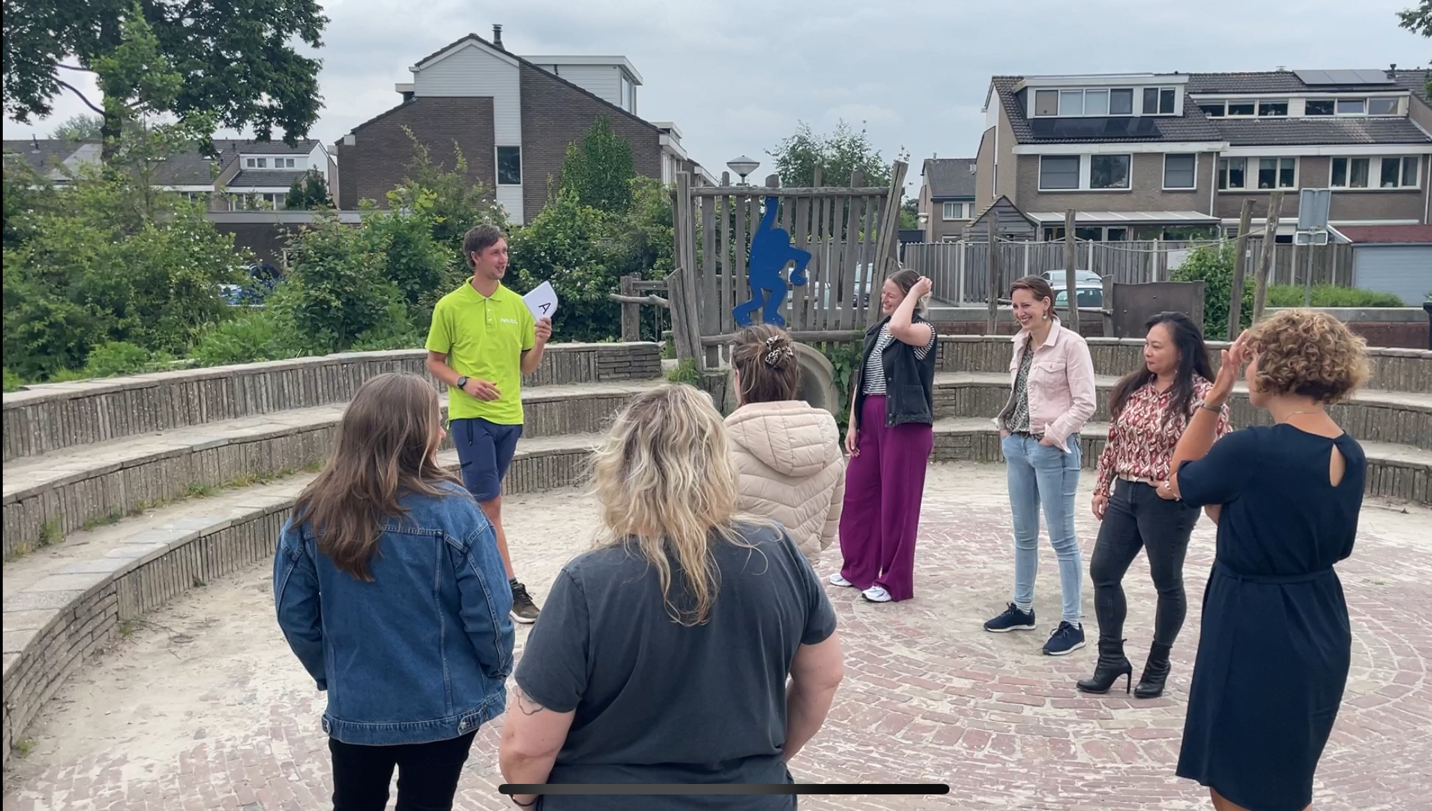 Groep mensen in gesprek op plein; man in groen shirt spreekt, omringd door luisteraars.