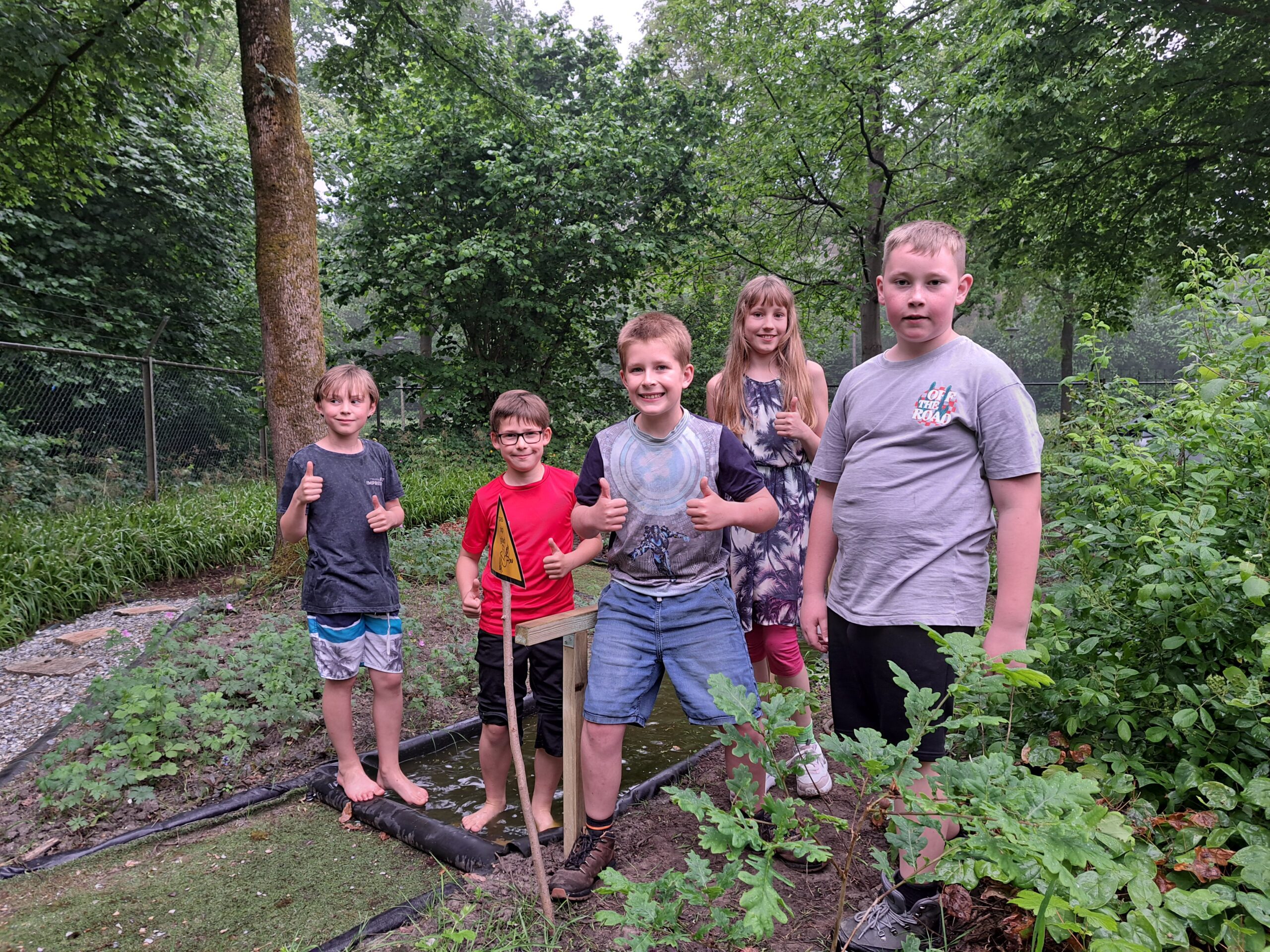 Vijf kinderen in een groene tuin, sommigen maken een duim omhoog gebaar.