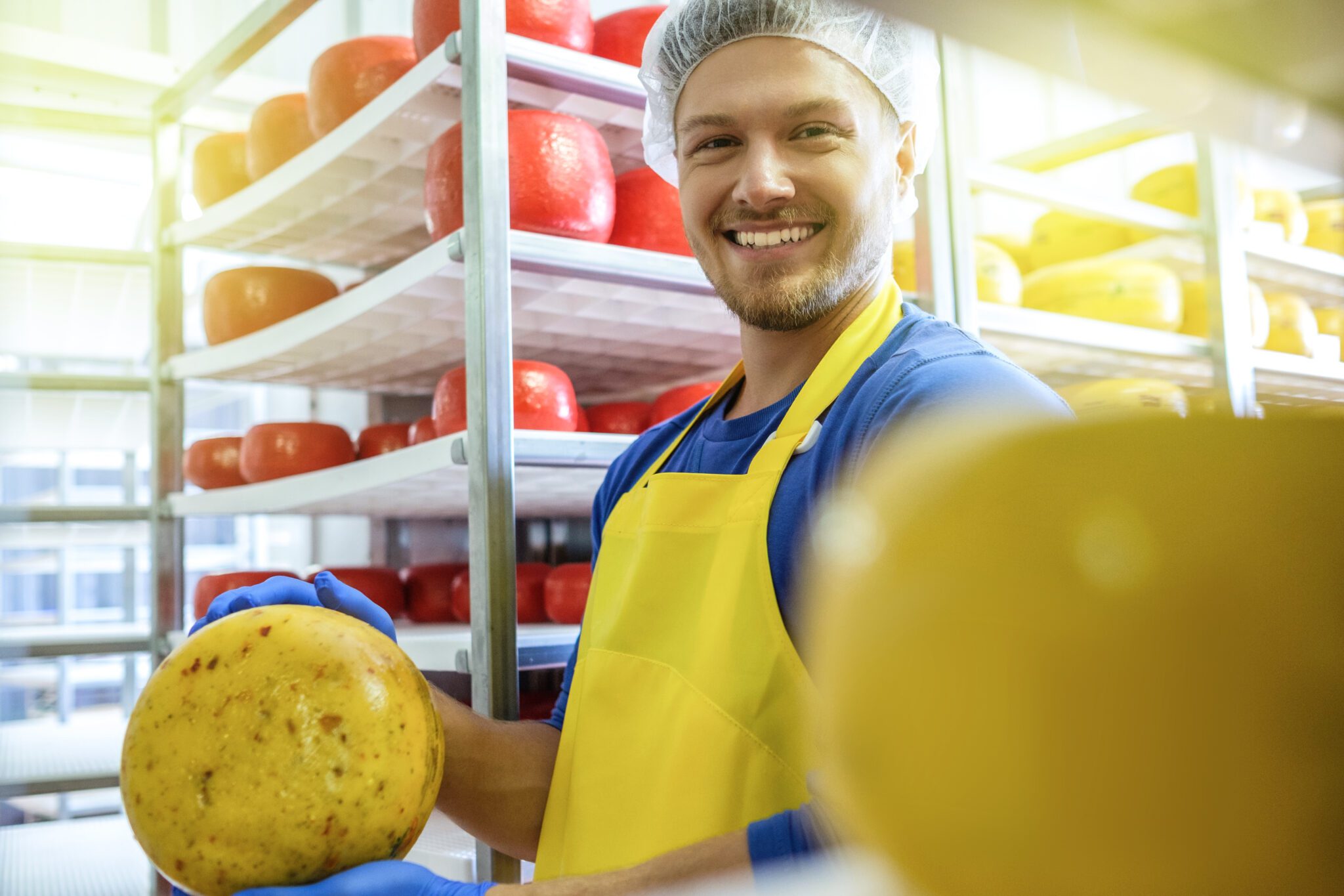 Man met gele schort toont kaas in een opslagruimte met planken vol kazen.