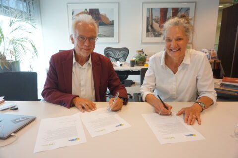 Twee mensen ondertekenen documenten aan een bureau in een kantooromgeving.