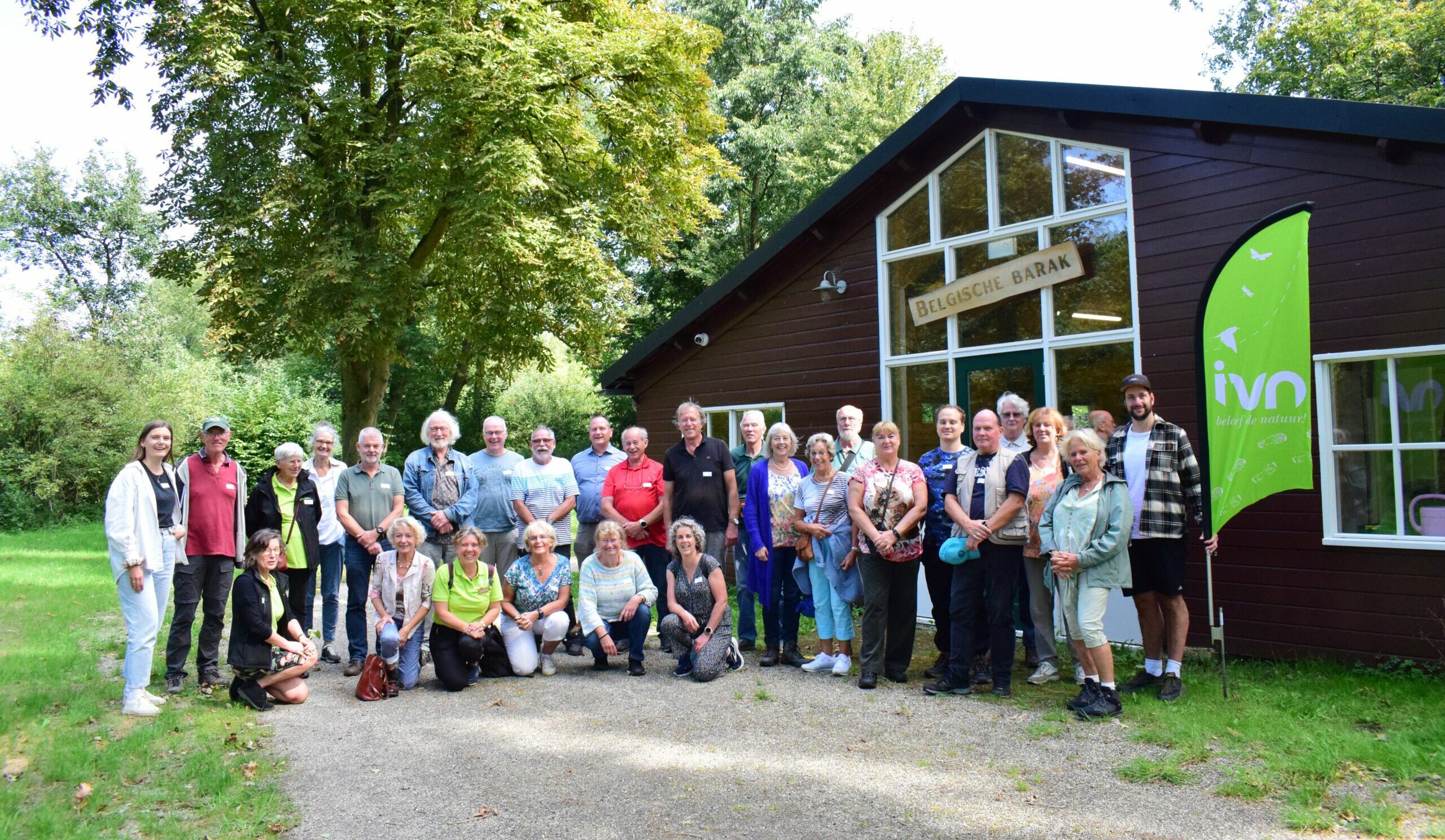 Groep mensen poseert voor een gebouw met "Belgische Barak," naast een groen IVN-vlag.