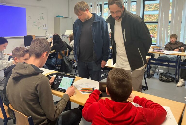 Studenten in een klaslokaal gebruiken een tablet voor een les; twee volwassenen kijken mee.