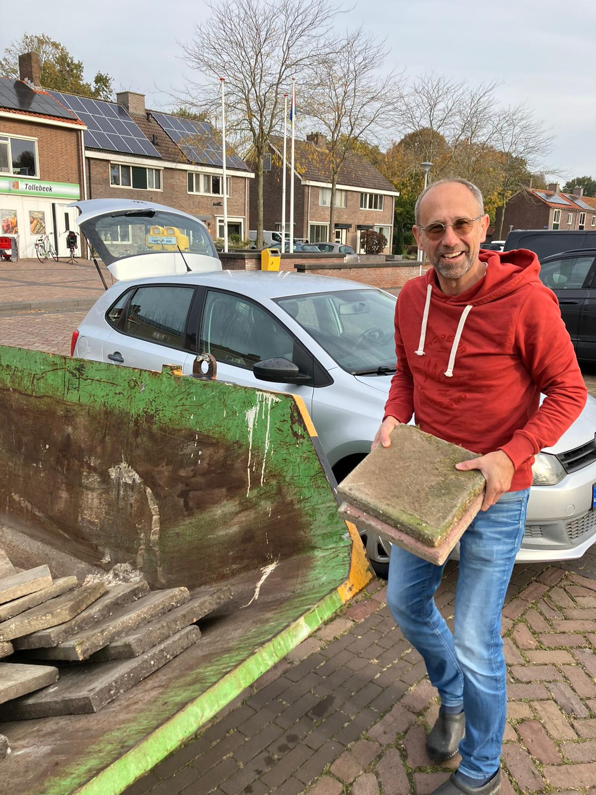 Man in rode trui laadt stoeptegels in een container naast een auto op een zonnige dag.