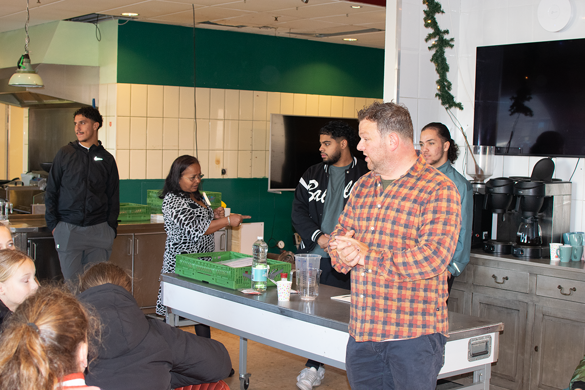 Man geeft presentatie aan groep mensen in keukenomgeving, met koffiezetapparaat op achtergrond.