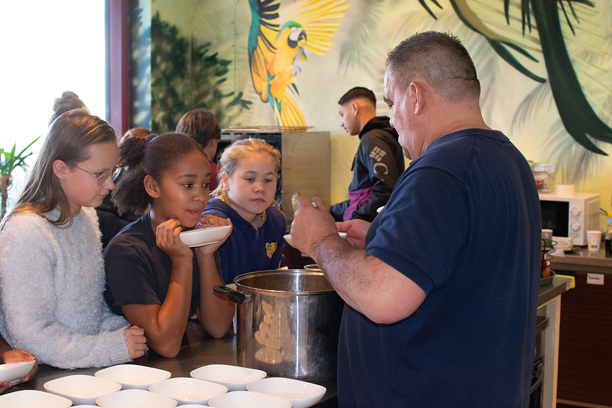 Man serveert soep aan drie kinderen bij een keuken met een tropisch muurschildering.