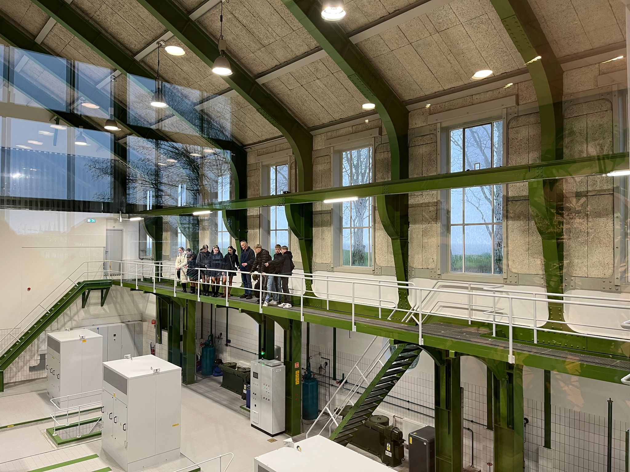 Industrieel interieur met groep op groene loopbrug, hoge ramen, apparatuur beneden.