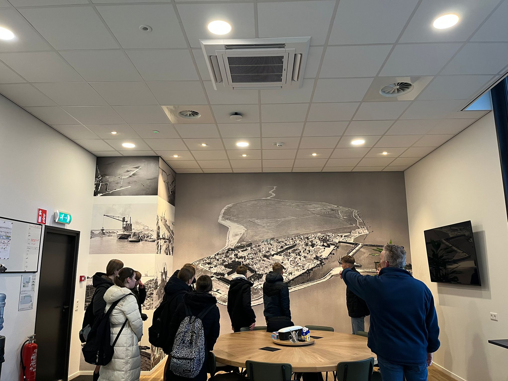 Groep mensen kijkt naar een grote muurkaart in een vergaderruimte met historische foto's.