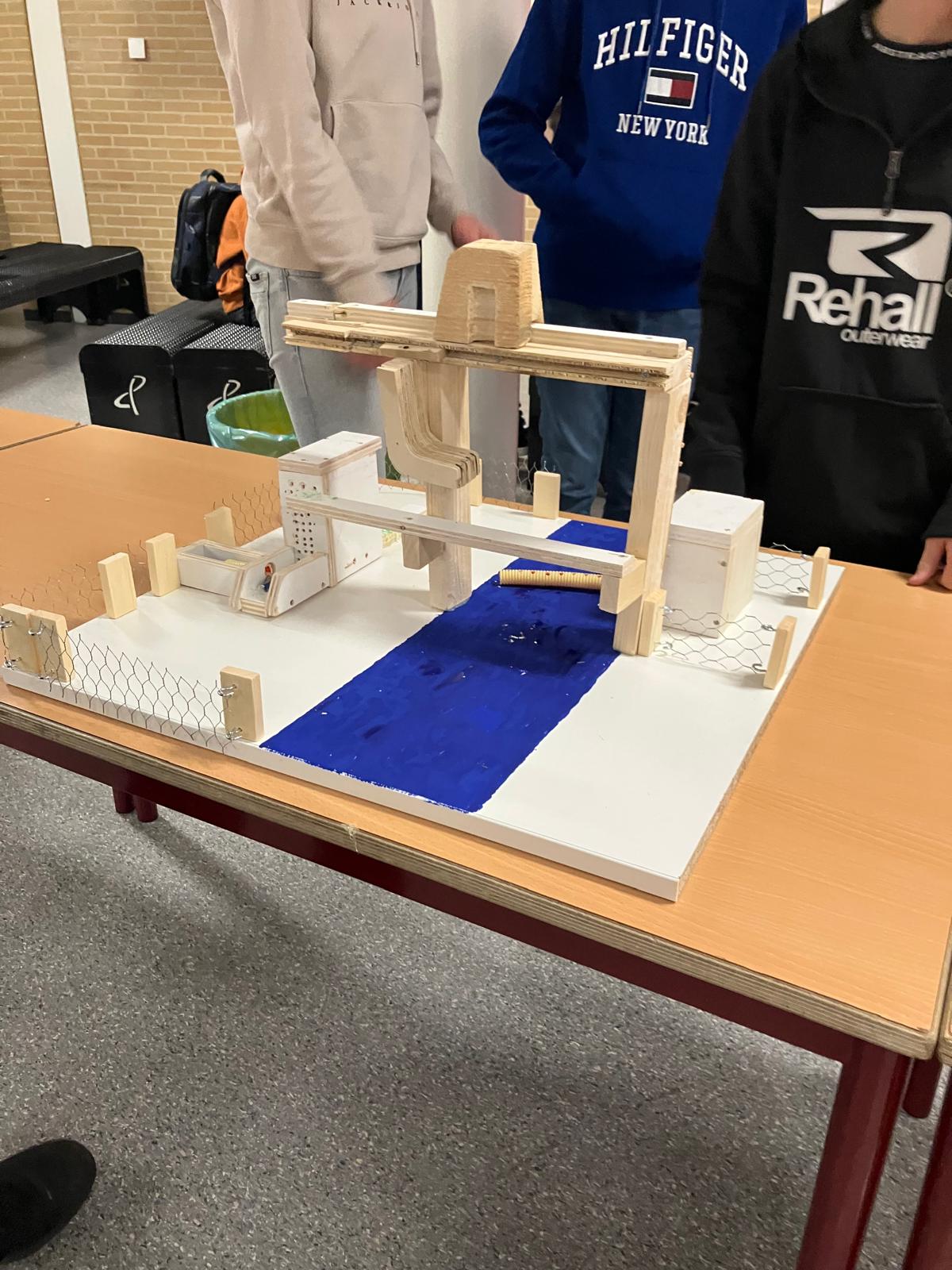 Model van een brug met houten constructie en blauwe rivier, omringd door studenten op een tafel.