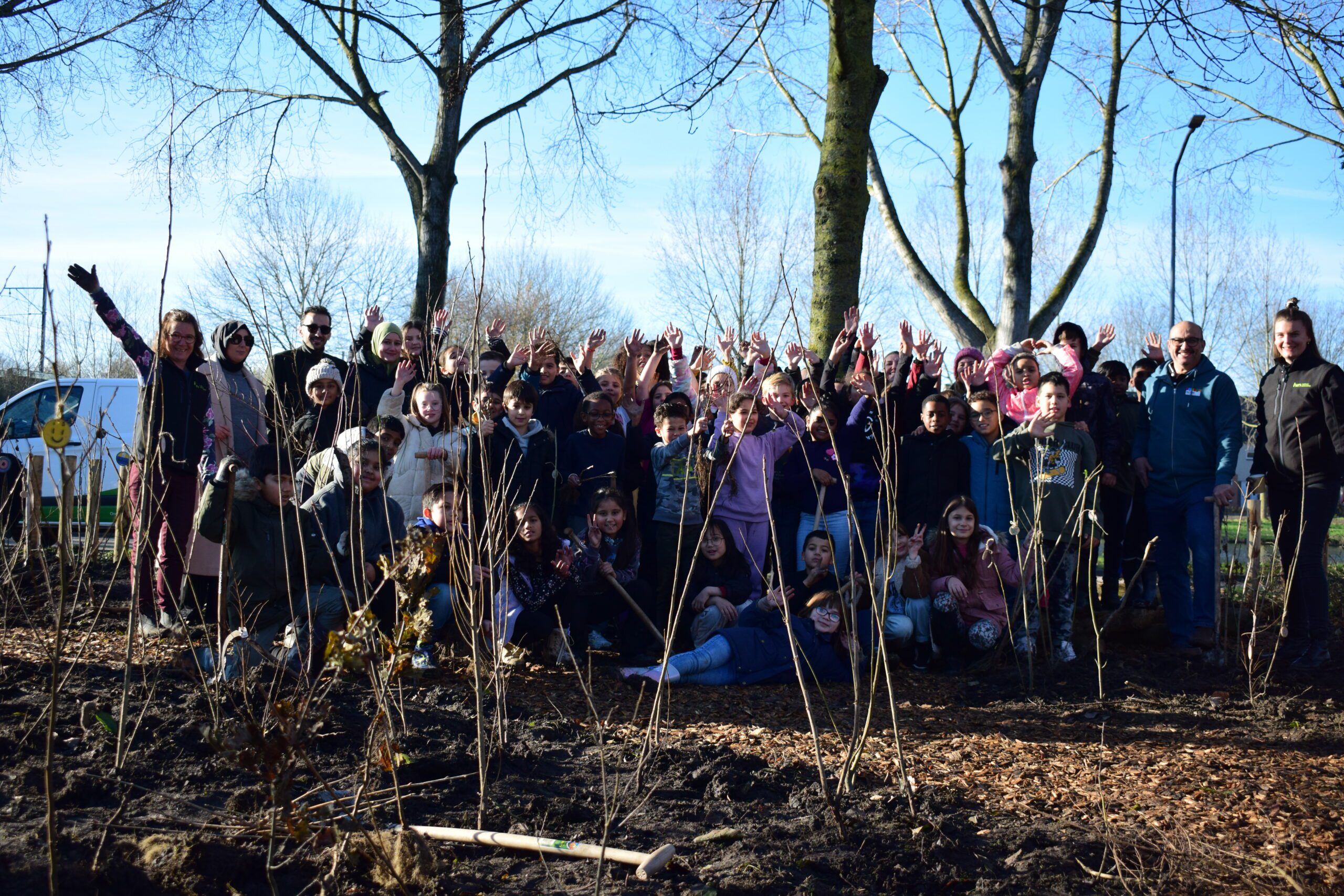 Groep mensen en kinderen poseert lachend bij jonge bomen in een park, bomen en blauwe lucht.