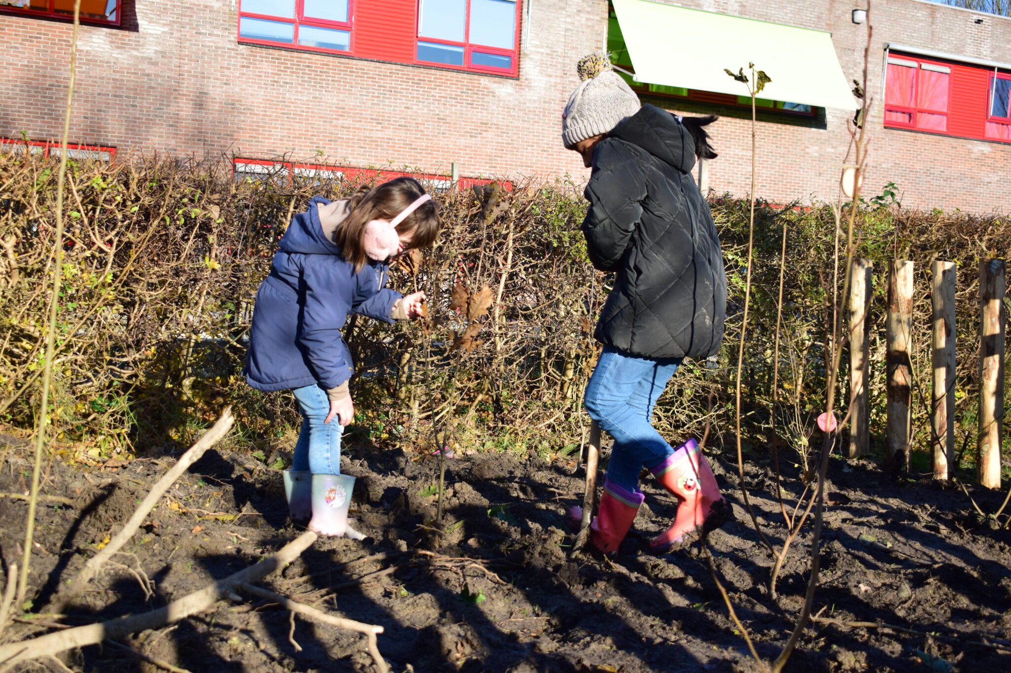 Twee kinderen planten bomen in een modderige tuin bij een bakstenen gebouw.