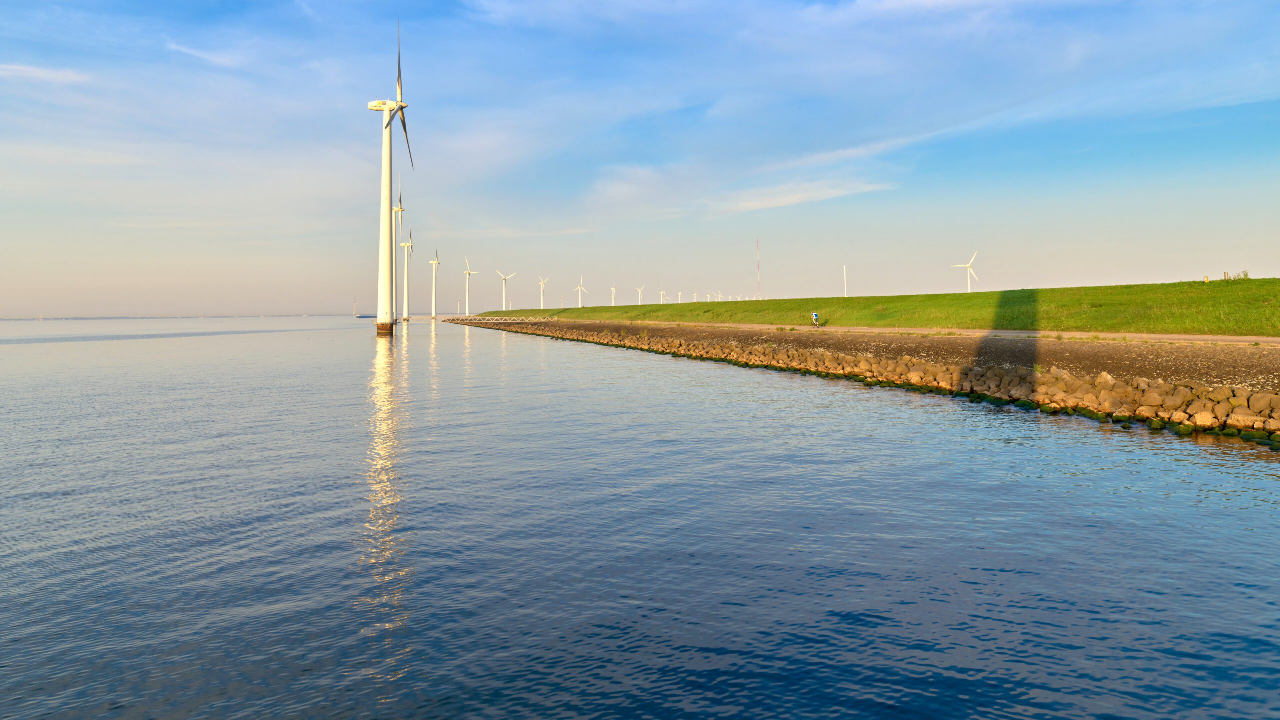 Rij windturbines langs waterkant onder een blauwe lucht met groene dijk en steenachtige oever.