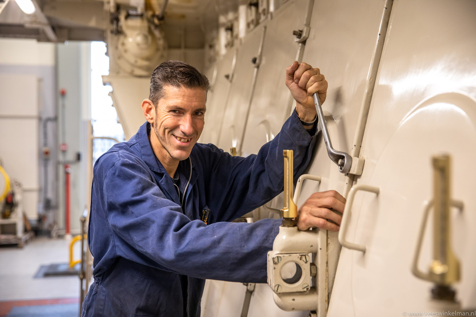 Een man in blauwe overall bedient industriële machines in een technische ruimte.