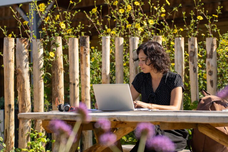 Vrouw werkt op laptop aan picknicktafel in zonnige tuin met bloemen en houten hek.