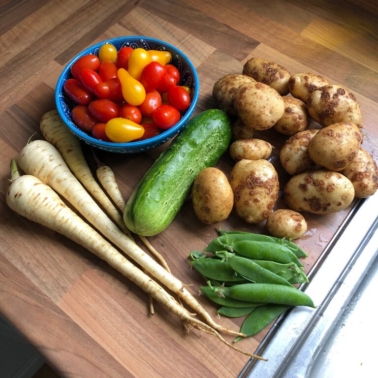 Verschillende groenten op een snijplank: een kom tomaten, pastinaak, komkommer, aardappelen en peultjes.