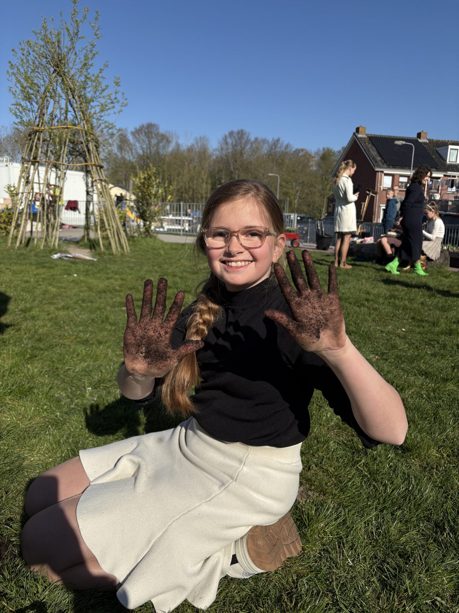 Meisje met modderige handen glimlacht op grasveld onder blauwe lucht, met groep mensen op de achtergrond.