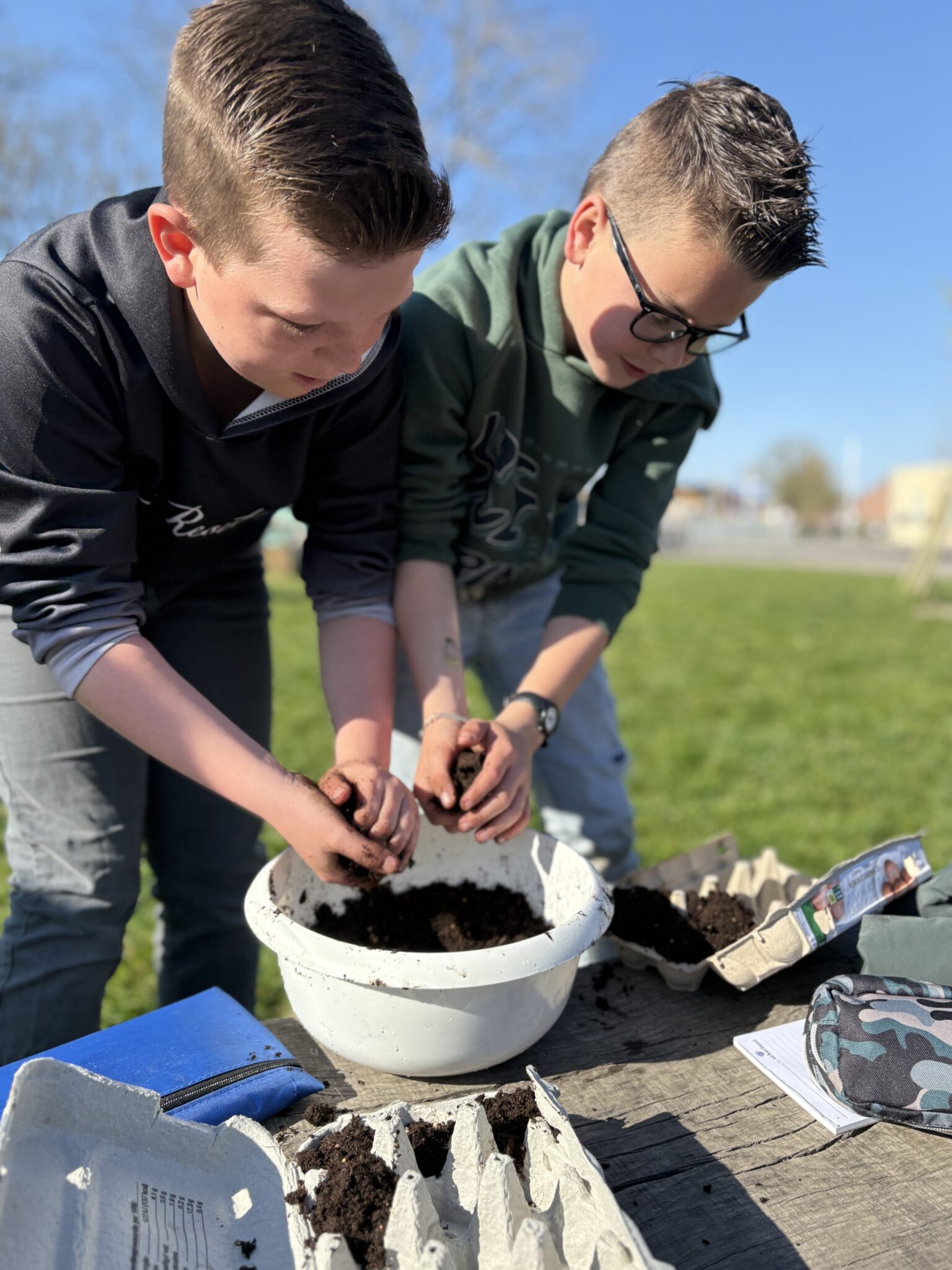 Twee kinderen planten zaden buiten, hun handen in potgrond boven een kom, met eierkartons.