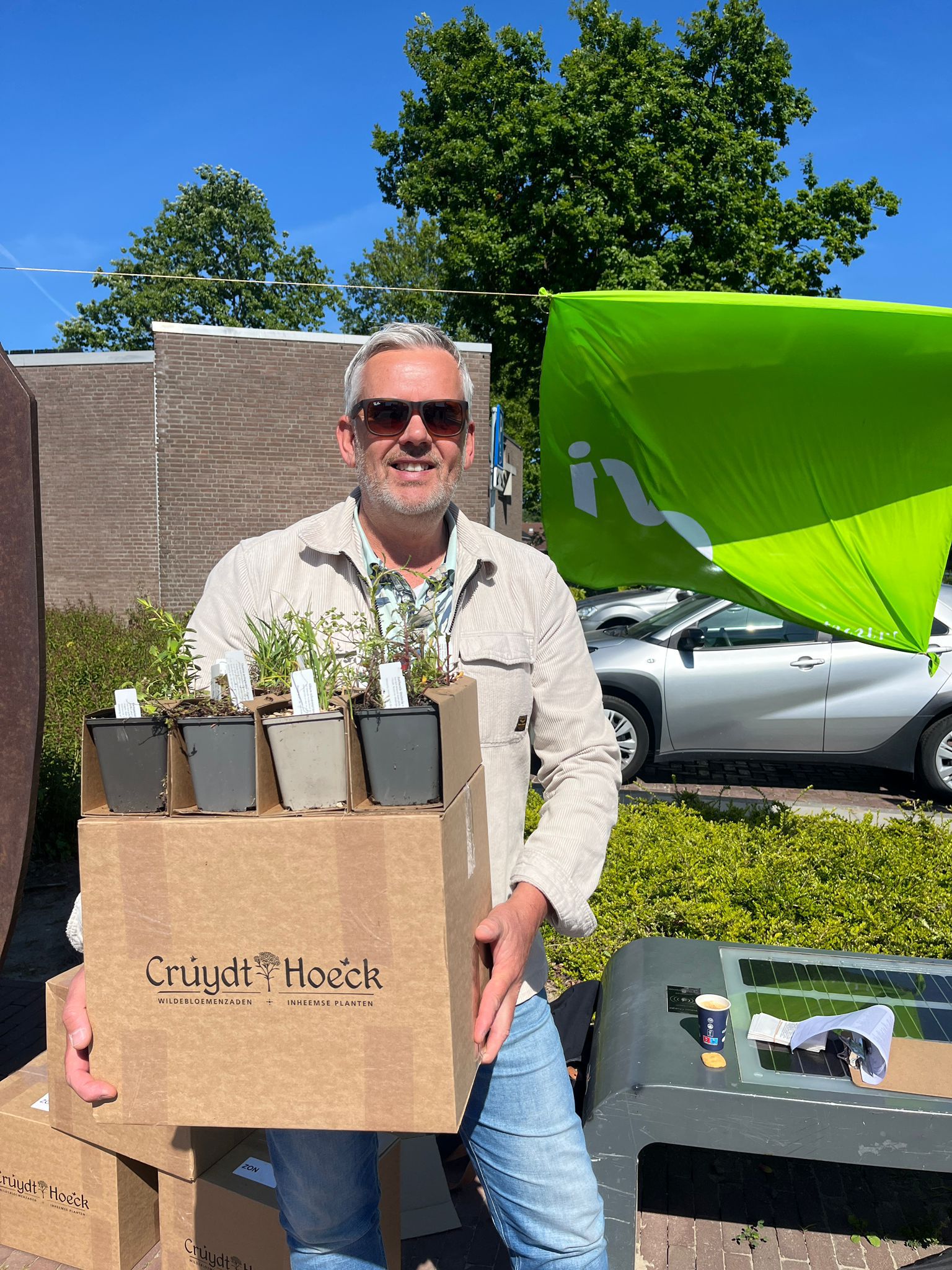 Persoon met zonnebril houdt een doos met plantjes vast, buiten bij een groene vlag en een auto.