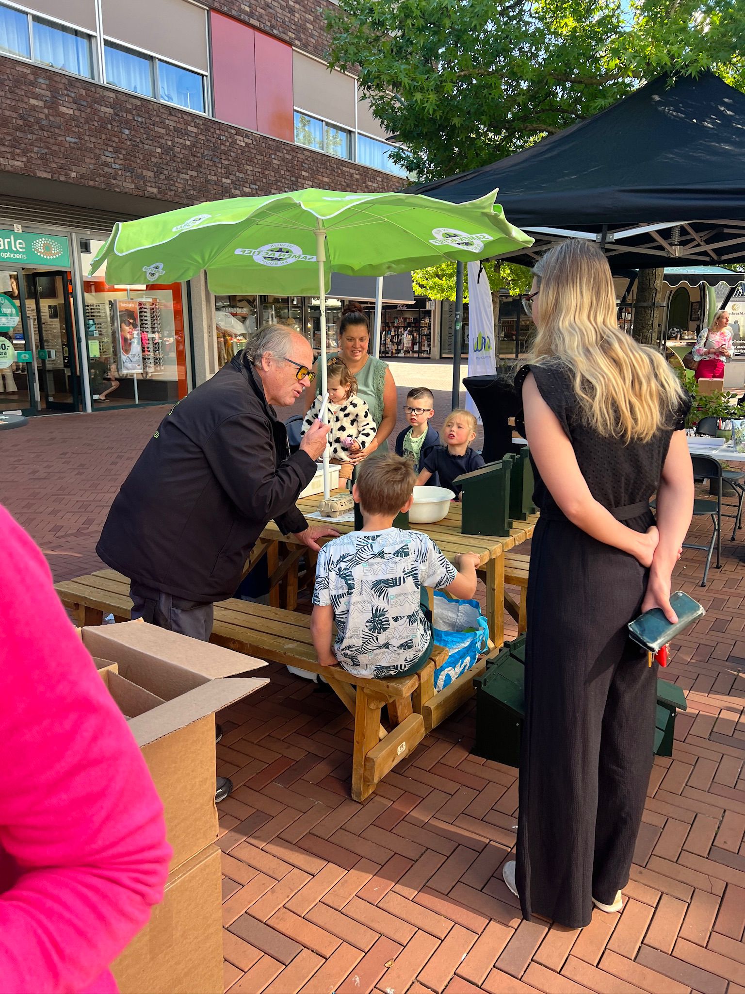 Een man spreekt kinderen aan een picknicktafel onder een groene parasol op een zonnig plein.
