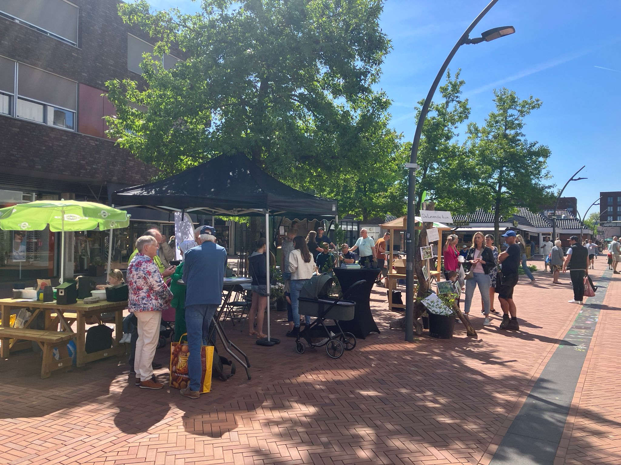 Mensen bezoeken een zonnige buitenmarkt met kraampjes en bomen op een plein.