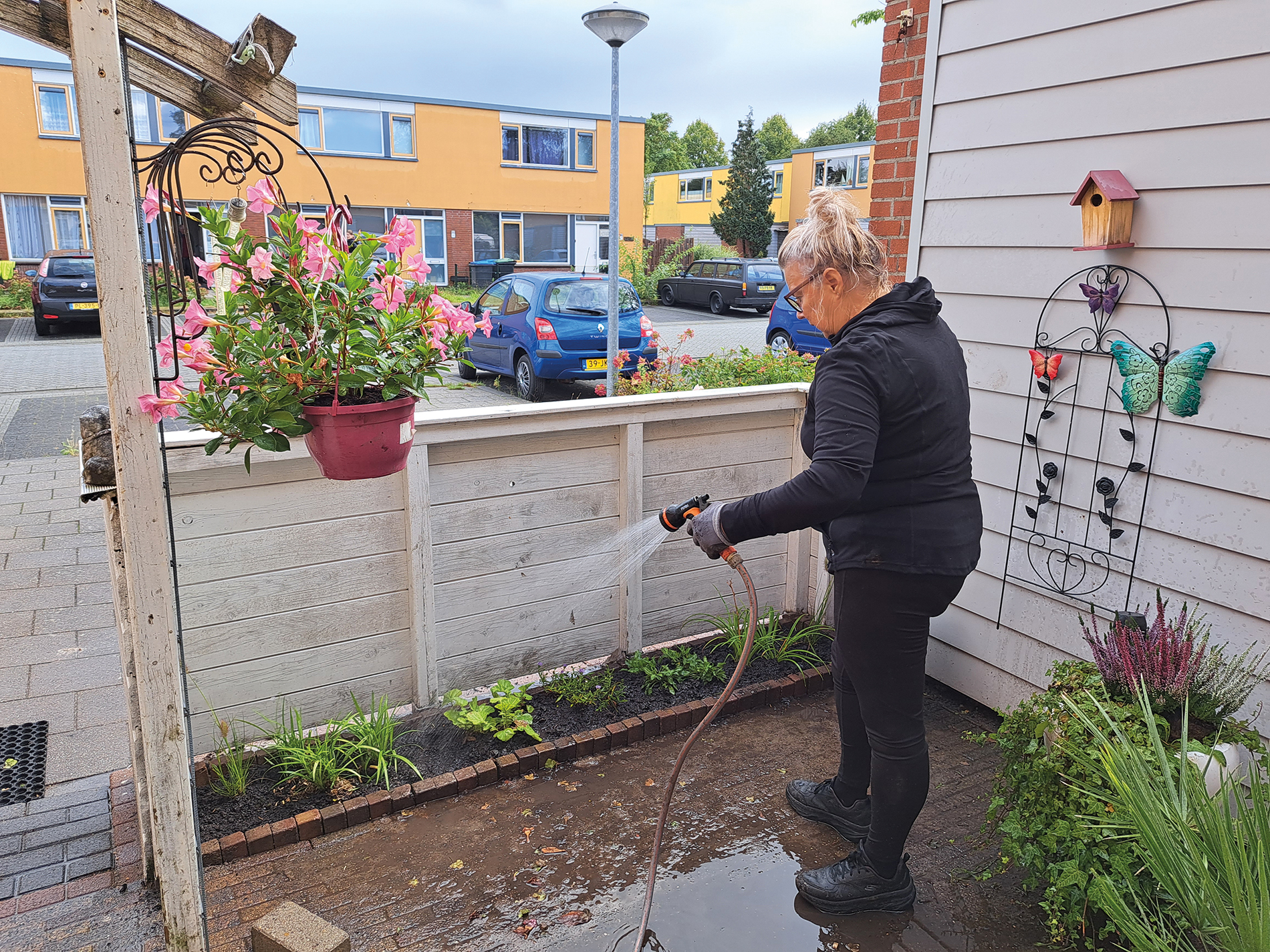 Een vrouw besproeit planten in een kleine tuin bij een huis met gele flats op de achtergrond.